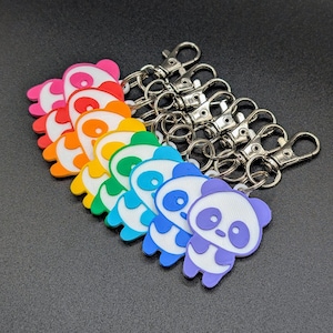 May include: A collection of colorful panda bear keychains. Each keychain features a different color scheme, including pink, red, orange, yellow, green, blue, and purple. The pandas have white faces and bodies, with black eyes and accents. Silver-tone metal clasps are attached.