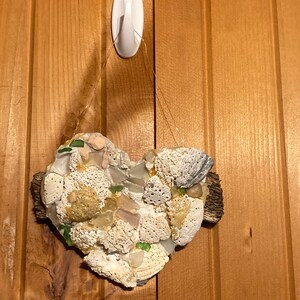 May include: A heart-shaped piece of driftwood decorated with seashells and sea glass. The heart is hanging from a hook on a wooden wall.