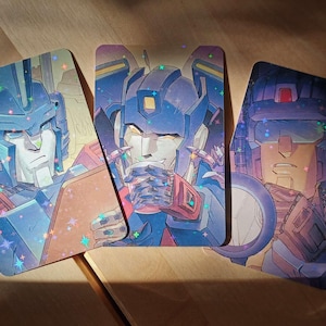 May include: Three illustrated cards featuring robots in blue, red, and white. The robots have detailed faces and are holding objects. The cards have a holographic effect, with sparkling stars. The background is a wooden surface.
