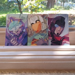 May include: Three character art prints depicting robots. The first print shows a purple and blue robot with a smiling expression. The second print features an orange and white robot. The third print shows a red and black robot.