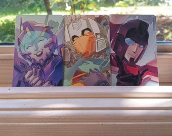 May include: Three colorful character art prints featuring robots. The first print has a purple and blue robot with a smiling face. The second print has an orange and white robot. The third print has a red and black robot.