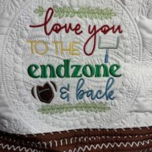 May include: White quilted throw blanket with a brown and white chevron pattern at the bottom. The top of the blanket features a circle with green leaves and the text "love you to the endzone & back" in a white, green, and brown font.