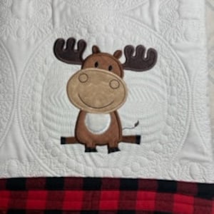 May include: A white quilted baby blanket with a brown moose applique. The blanket has a red and black buffalo plaid trim.