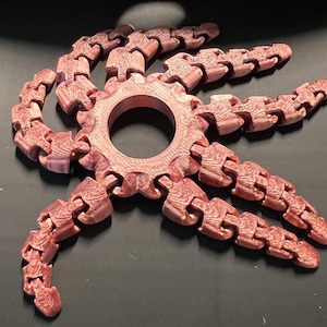 May include: A reddish-brown, articulated fidget toy resembling an octopus or gear. The central ring has a large hole, and eight flexible arms extend outward. The toy has a wood-grain texture, and is designed to be manipulated.