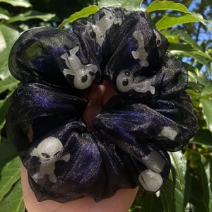 May include: Black scrunchie with a sheer overlay, featuring a repeating pattern of small, white ghost figures. The scrunchie is gathered and appears to be made of a soft, lightweight fabric. The background is a natural outdoor setting.
