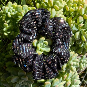 May include: A black scrunchie with a shimmering, sequined texture. The fabric is covered in small, rectangular sequins that catch the light, creating a sparkling effect. The scrunchie is set against a backdrop of green succulent plants.