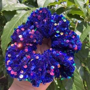 May include: A vibrant blue scrunchie, densely covered in iridescent sequins. The hair accessory is made of a soft, plush material and is designed to hold hair in place. The sequins catch the light, creating a sparkling effect. The scrunchie is a stylish hair accessory.