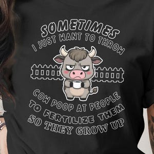 May include: Black t-shirt featuring a grumpy cartoon cow holding a cup, set against a fence graphic. The text says "Sometimes I just want to throw cow poop at people to fertilize them so they grow up."