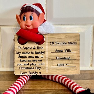 May include: A festive Christmas elf doll with red and white striped legs and black shoes. The elf holds a wooden sign with text: "To Sophie & Beth, My name is Buddy! Santa sent me to keep an eye on you and play until Christmas Day. Love Buddy x".
