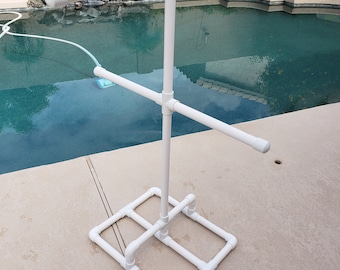 May include: White PVC pipe pool towel rack with four horizontal bars and a square base. The rack is standing on a concrete patio next to a swimming pool.