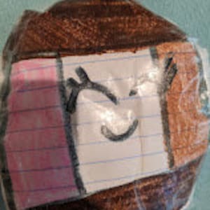 Op de afbeelding: Een veelkleurige, ronde papier-maché bal met een knipperend gezicht erop getekend. De bal is versierd met roze, bruin en wit papier.