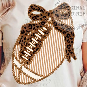 Può includere: T-shirt color crema con un design a pallone da football americano, ornato da un fiocco leopardato e dettagli. Il pallone presenta strisce verticali e una catena di perline. Il testo "ORIGINAL DESIGNER" è visibile.