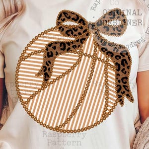 以下が含まれることがあります： ヒョウ柄のリボンが付いたバスケットボールのデザインのクリーム色のTシャツ。バスケットボールには縦縞とビーズの輪郭があります。「ORIGINAL DESIGNER」の文字がシャツに表示されています。