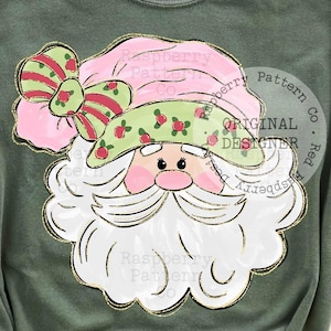 May include: Olive green t-shirt featuring a Santa Claus graphic. The Santa design includes a pink hat with a green bow, a white beard, and a gold outline. The text "Raspberry Pattern Co. Original Designer" is printed on the shirt.