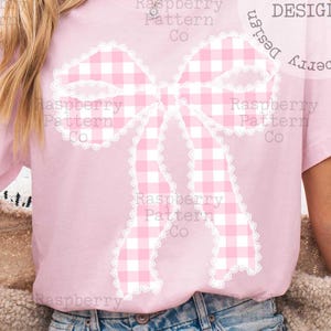 May include: A light pink t-shirt featuring a large bow design. The bow has a gingham pattern in pink and white, outlined with a white lace trim. The text "Raspberry Pattern Co" is printed on the shirt.