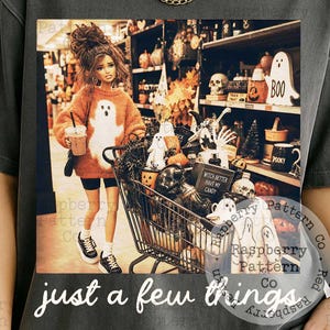 May include: A charcoal gray short-sleeve t-shirt featuring a graphic print of a doll in a store aisle with a shopping cart filled with Halloween decor. The text "just a few things" is printed in white below the image on the front of the shirt.