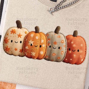 May include: Beige t-shirt with a row of four smiling pumpkin appliqués in orange, tan, and brown. Each pumpkin has a different floral pattern. A silver chain necklace rests on the shirt.