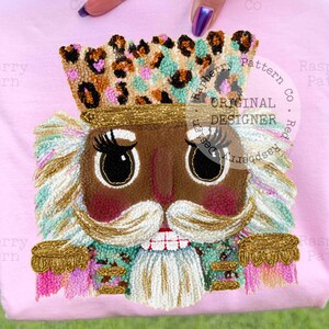May include: A light pink t-shirt featuring an embroidered nutcracker face with a leopard print crown, gold accents, and a white mustache. The design includes the text "ORIGINAL DESIGNER".