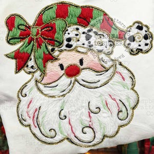 May include: Embroidered Santa Claus design on a white pillowcase. Santa features a red nose, rosy cheeks, and a white beard with gold trim. His hat is green and red striped with a matching bow. The design includes the text "ORIGINAL DESIGNER".