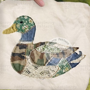May include: A decorative duck applique, featuring a patchwork design with denim, camouflage, floral, and lace fabrics. The duck has a green floral head, a yellow beak, and blue denim accents. The design is set on a cream-colored background.