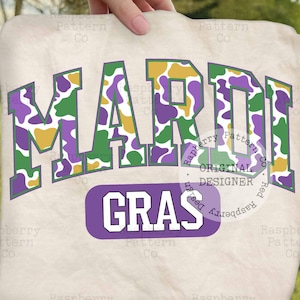 Peut inclure: Un sweat-shirt crème avec l'inscription "MARDI GRAS" en grandes lettres colorées. Les lettres présentent un motif camouflage vert, violet et or. En dessous, le mot "GRAS" est dans un rectangle violet.