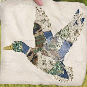 May include: A square, cream-coloured fabric panel with a patchwork duck design. The duck is made from diverse fabrics like camouflage, denim, and lace, with the text "ORIGINAL DESIGNER". The duck has a green head and yellow beak.