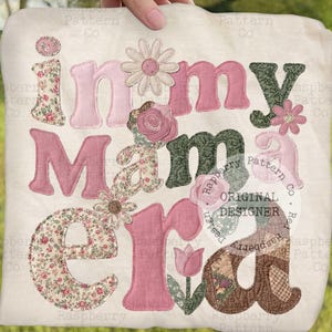 Puede incluir: Un bolso tote crema con la frase "in my Mama era" en letras aplicadas rosas, florales y verdes. El diseño incluye detalles florales y de margaritas. El bolso está sostenido por una mano.