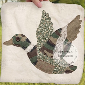 May include: A cream-colored square fabric with a patchwork duck design. The duck features camouflage, floral, and lace details, with a button eye. The words "ORIGINAL DESIGNER" are visible.