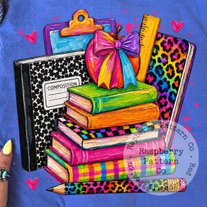 May include: A periwinkle blue t-shirt featuring a colorful graphic of a stack of books, a composition notebook, a clipboard, a ruler, and a pencil. The design includes a bright apple with a rainbow bow, leopard print accents, and small hearts.