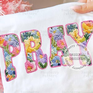 Könnte beinhalten: Weißes T-Shirt mit dem Wort "PRAY", das mit farbenfrohen Blumenmustern bestickt ist. Die Buchstaben sind pink umrandet und bilden ein lebendiges Design. Die Stickerei zeigt eine Vielzahl von Blumen.