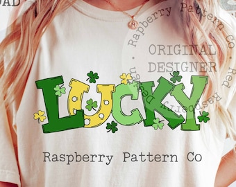 May include: A white t-shirt with a green, yellow, and gold graphic that says "LUCKY" with shamrocks and a horseshoe. The text "Raspberry Pattern Co." is printed below the graphic.