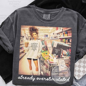 May include: A charcoal gray crewneck t-shirt featuring a graphic print of a person in a store aisle with a shopping cart full of school supplies. The person wears a sweatshirt with the text Teacher Fueled By Iced Coffee & Pure Chaos.