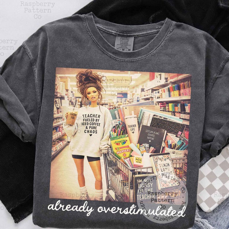 May include: A charcoal gray crewneck t-shirt featuring a graphic print of a person in a store aisle with a shopping cart full of school supplies. The person wears a sweatshirt with the text Teacher Fueled By Iced Coffee & Pure Chaos.