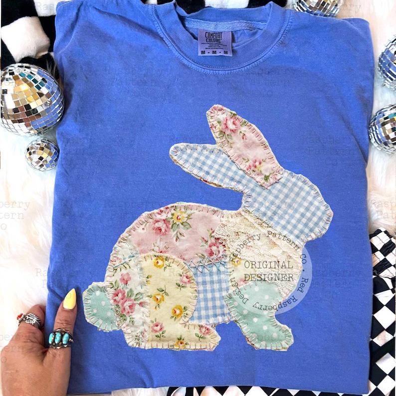 May include: A blue Comfort Colors t-shirt featuring a patchwork bunny design. The bunny is made of floral, gingham, and lace fabrics in pastel colors. The shirt has a crew neck and short sleeves.