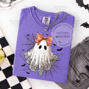 K&ouml;nnte beinhalten: Ein lila T-Shirt mit einem Geistermotiv, Schleife, Spinnweben und Flederm&auml;usen. Der Geist ist wei&szlig; mit schwarzer Spitze und Augen. Das T-Shirt hat einen V-Ausschnitt und eine silberne Halskette. Der Text "ORIGINAL DESIGNER" ist sichtbar.