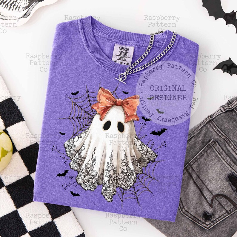 K&ouml;nnte beinhalten: Ein lila T-Shirt mit einem Geistermotiv, Schleife, Spinnweben und Flederm&auml;usen. Der Geist ist wei&szlig; mit schwarzer Spitze und Augen. Das T-Shirt hat einen V-Ausschnitt und eine silberne Halskette. Der Text "ORIGINAL DESIGNER" ist sichtbar.