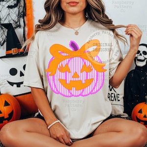 以下が含まれることがあります： ハロウィーンをテーマにしたカボチャのデザインが施されたクリーム色のTシャツ。カボチャはピンクと白のストライプ模様、オレンジ色のリボン、ジャック・オー・ランタンの顔をしています。「Raspberry Pattern Co」の文字が見えます。ハロウィーンのデザイン。