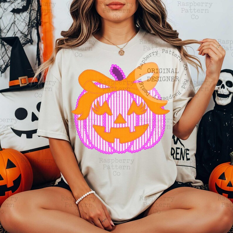 以下が含まれることがあります： ハロウィーンをテーマにしたカボチャのデザインが施されたクリーム色のTシャツ。カボチャはピンクと白のストライプ模様、オレンジ色のリボン、ジャック・オー・ランタンの顔をしています。「Raspberry Pattern Co」の文字が見えます。ハロウィーンのデザイン。