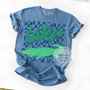 May include: Blue t-shirt featuring a green alligator illustration and the word "Later" in green. The design includes a checkered pattern and green stars. A casual wear item.