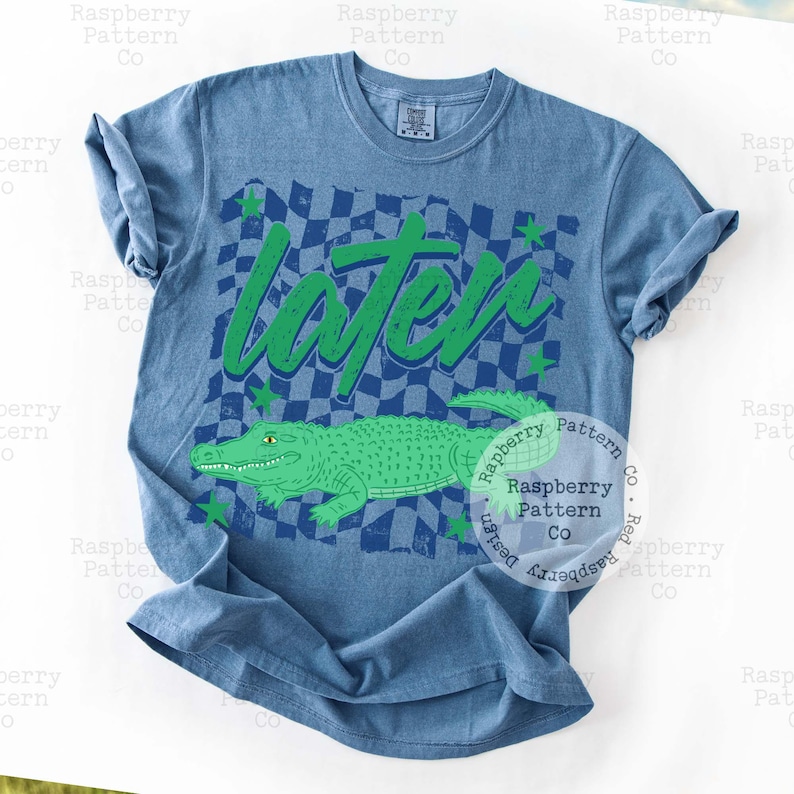 May include: Blue t-shirt featuring a green alligator illustration and the word "Later" in green. The design includes a checkered pattern and green stars. A casual wear item.