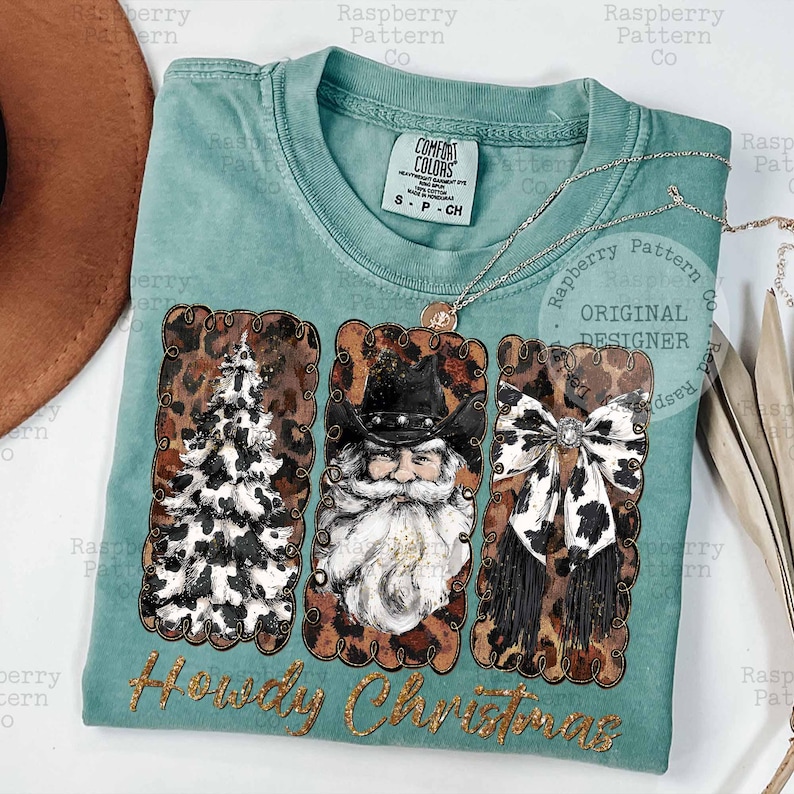 May include: A seafoam green t-shirt with a festive design. The shirt features three rectangular panels with a Christmas tree, Santa Claus, and a bow, all with a cowhide pattern. The words "Howdy Christmas" are written in gold script.