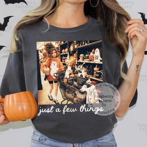 May include: A charcoal gray t-shirt featuring a graphic print of a doll wearing an orange sweater with a ghost design, pushing a shopping cart filled with Halloween decor. The text "just a few things" is printed in white below the graphic.