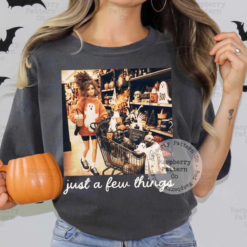 May include: A charcoal gray t-shirt featuring a graphic print of a doll wearing an orange sweater with a ghost design, pushing a shopping cart filled with Halloween decor. The text "just a few things" is printed in white below the graphic.