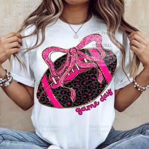 May include: White t-shirt featuring a pink and black football design with a bow and the words "game day". The football has a leopard print pattern and pink laces. The design is outlined in gold.