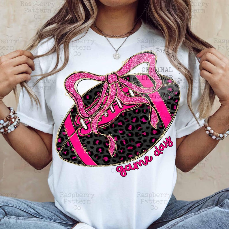 May include: White t-shirt featuring a pink and black football design with a bow and the words "game day". The football has a leopard print pattern and pink laces. The design is outlined in gold.