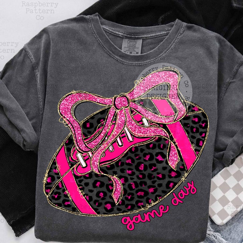 May include: Dark gray sweatshirt with a pink glitter football and bow design. The football has a leopard print pattern and pink stripes. The words "game day" are written in pink script.