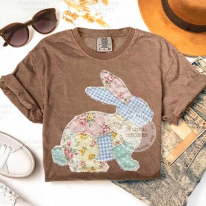 May include: A brown Comfort Colors t-shirt featuring a patchwork bunny design. The bunny is made of various floral and gingham fabrics in pastel colors. The shirt has rolled-up sleeves and a crew neck. The text "ORIGINAL DESIGNER" is visible.