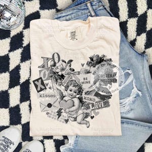 May include: Cream-colored t-shirt featuring a vintage-style graphic of a cherub holding a heart, surrounded by floral elements, the words "XO," "kisses," and "LOVE." The shirt is laid on top of denim jeans and a black and white checkered surface.