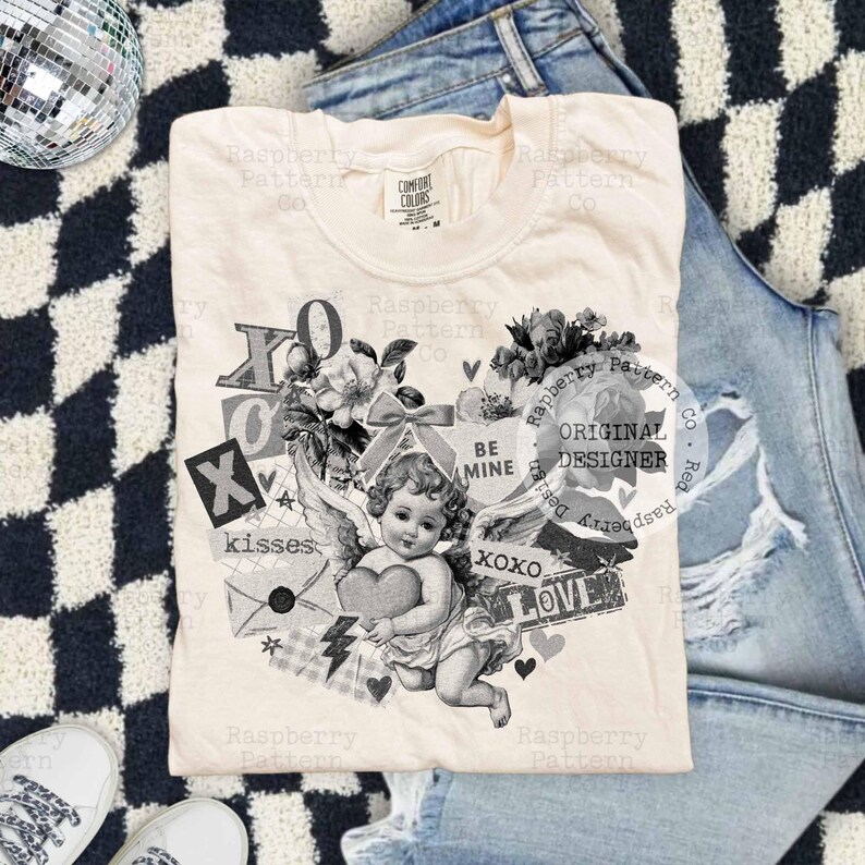 May include: Cream-colored t-shirt featuring a vintage-style graphic of a cherub holding a heart, surrounded by floral elements, the words "XO," "kisses," and "LOVE." The shirt is laid on top of denim jeans and a black and white checkered surface.