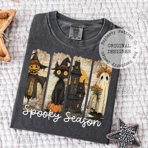 May include: Dark gray t-shirt featuring a Halloween-themed graphic. The design includes a scarecrow, black cat, haunted house, and ghost, with the words "Spooky Season" in white script. The shirt has a crew neck and short sleeves.
